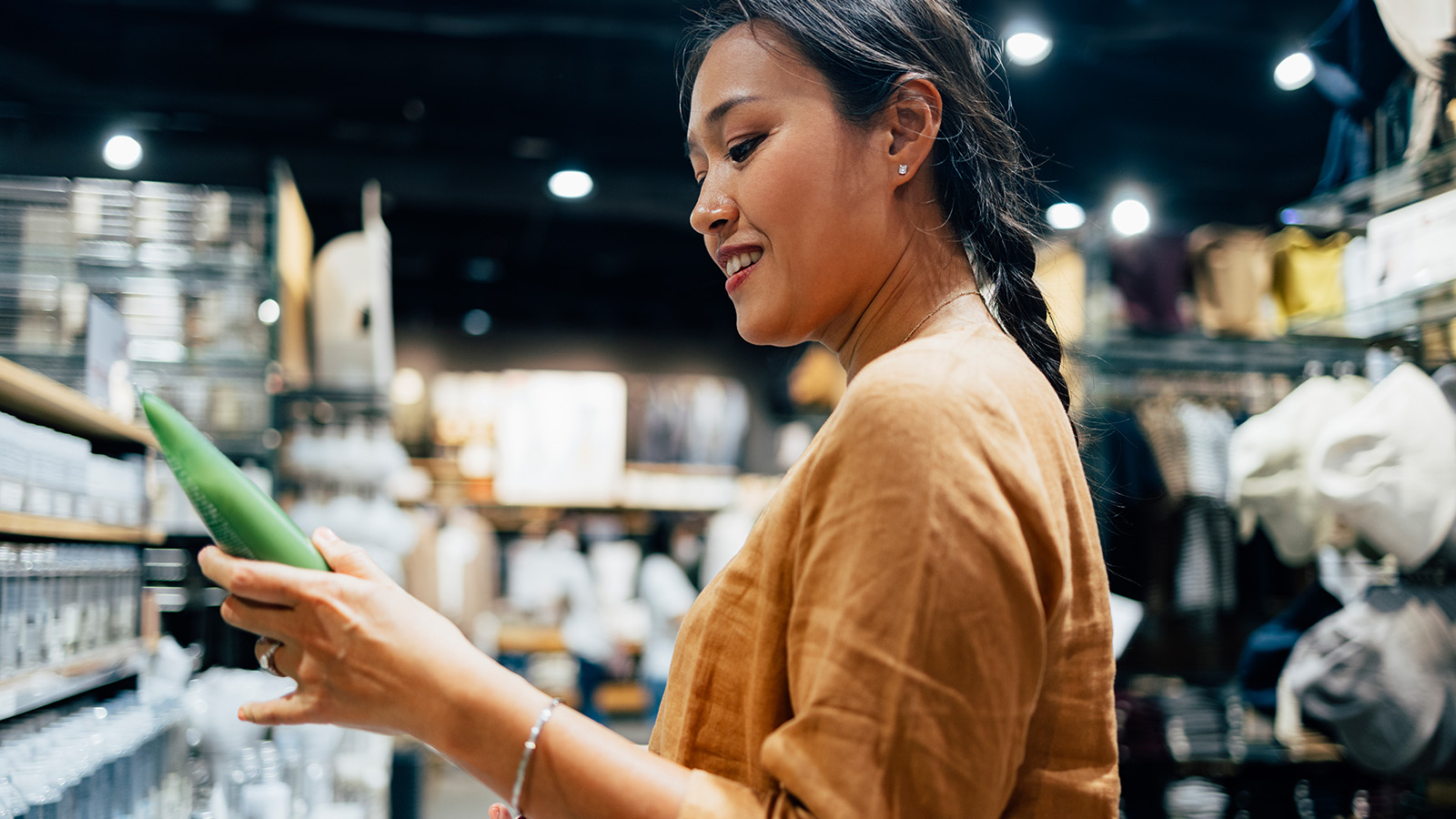 A side view of a smiling Asian female at a store holding a tube of some cosmetic product and deciding whether to buy it or not