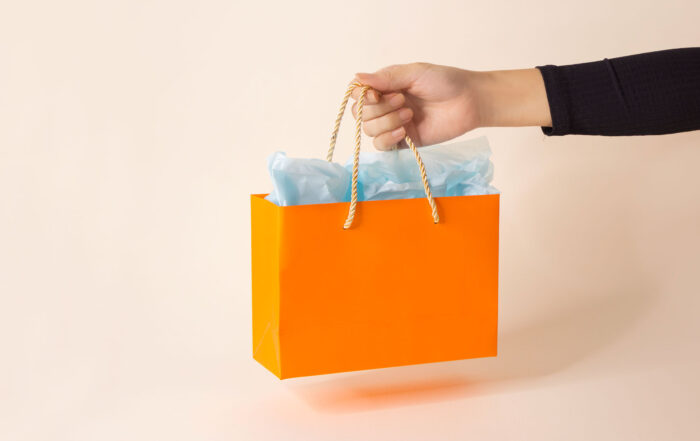 One women's hand holding one orange colour shopping paper bag on pink background.