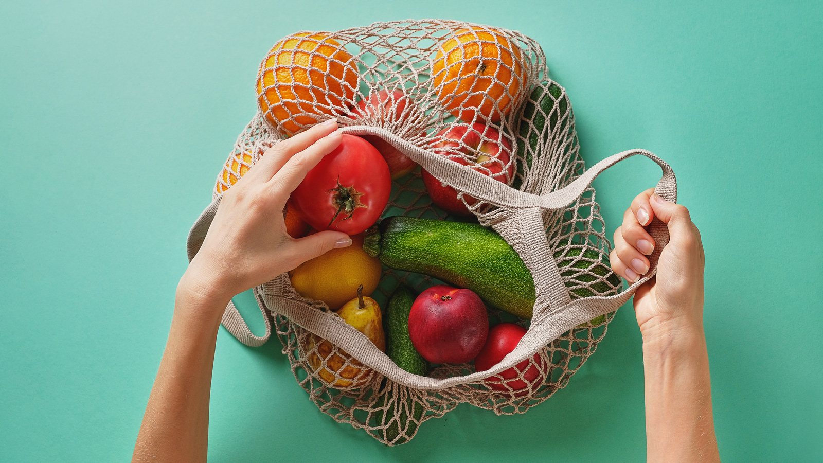 Fresh juicy fruits and vegetables, products in a reusable shopping bag. A girl or woman takes or lays out products from a string bag made from recycled materials on a Green Pastel background. Vegetarianism, Veganism. No plastic.