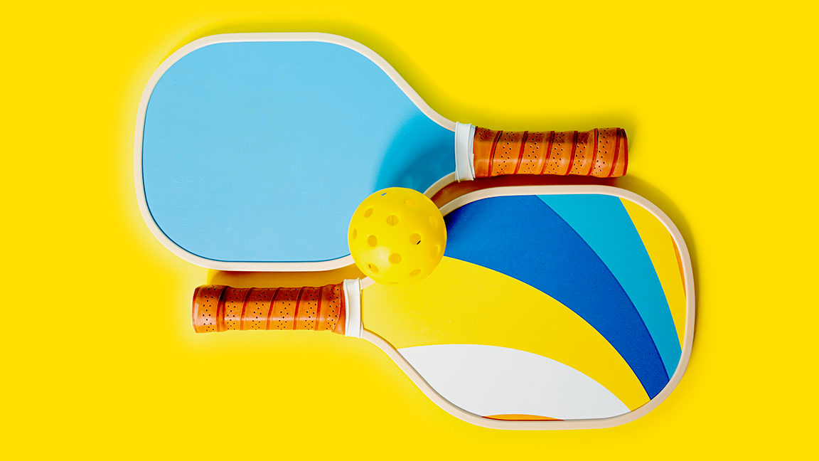 Photograph of two pickleball rackets and a ball on a yellow background. The rackets are blue and yellow. Pickle ball concept. Equipment for playing pickleball.