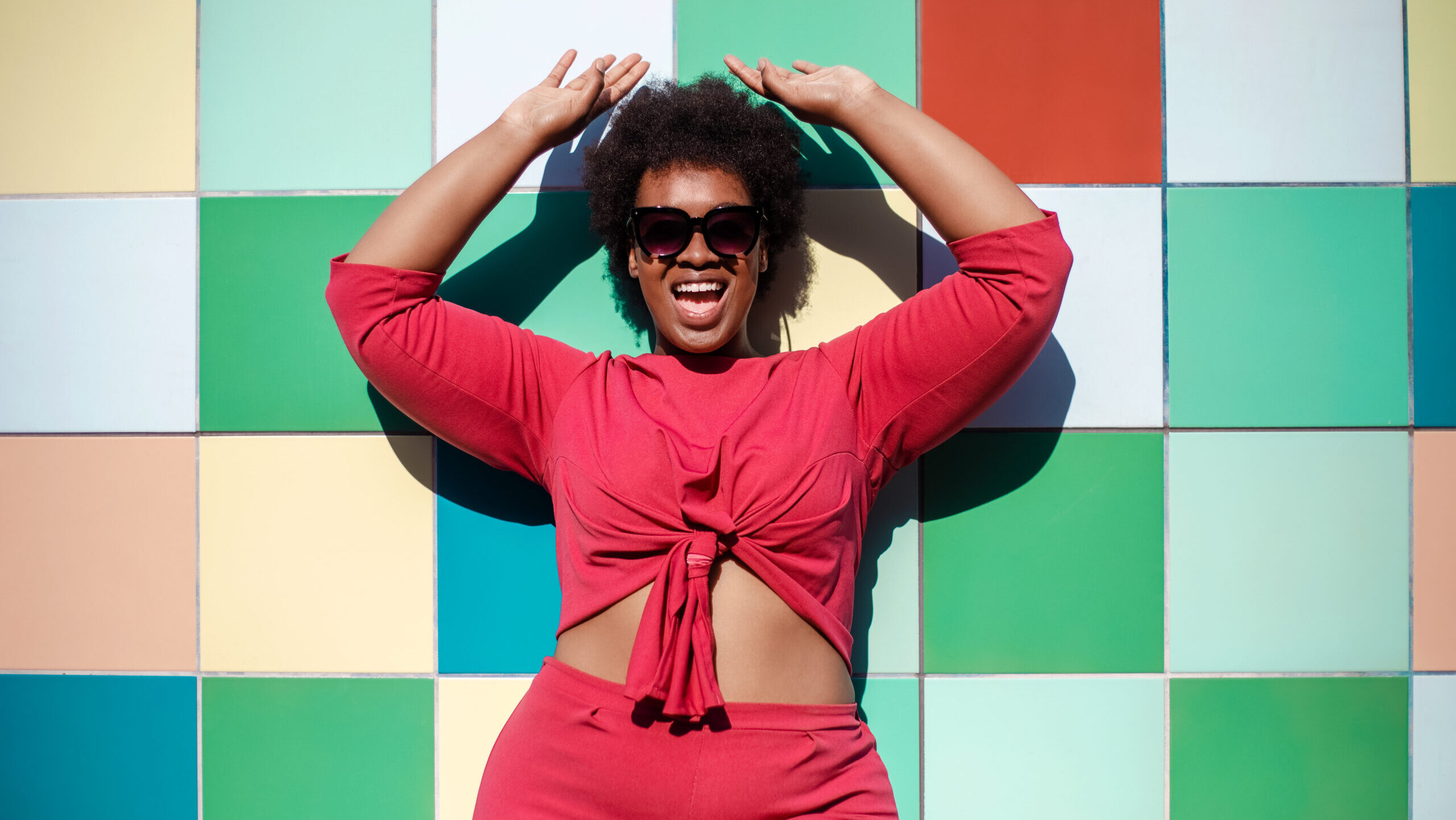 Young woman in stylish casuals and sunglasses looking excited against multicolored tiled wall. African american female looking at camera and smiling.
