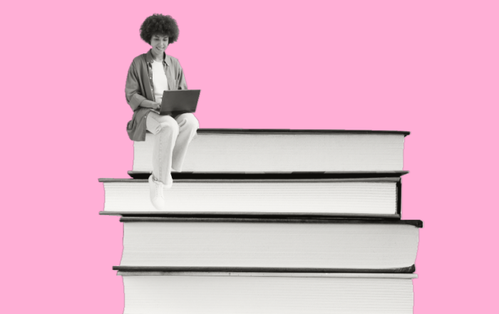 A woman sitting on top of a stack of closed books while browsing on their laptop computer in front of a pink background.