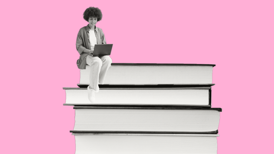 A woman sitting on top of a stack of closed books while browsing on their laptop computer in front of a pink background.